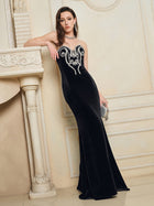 A woman in the Social Queen Hand-Applied Silver Crystal Baroque Velvet Gown, featuring a strapless fitted silhouette and white embroidery, stands indoors by a decorative fireplace, holding a small silver clutch.