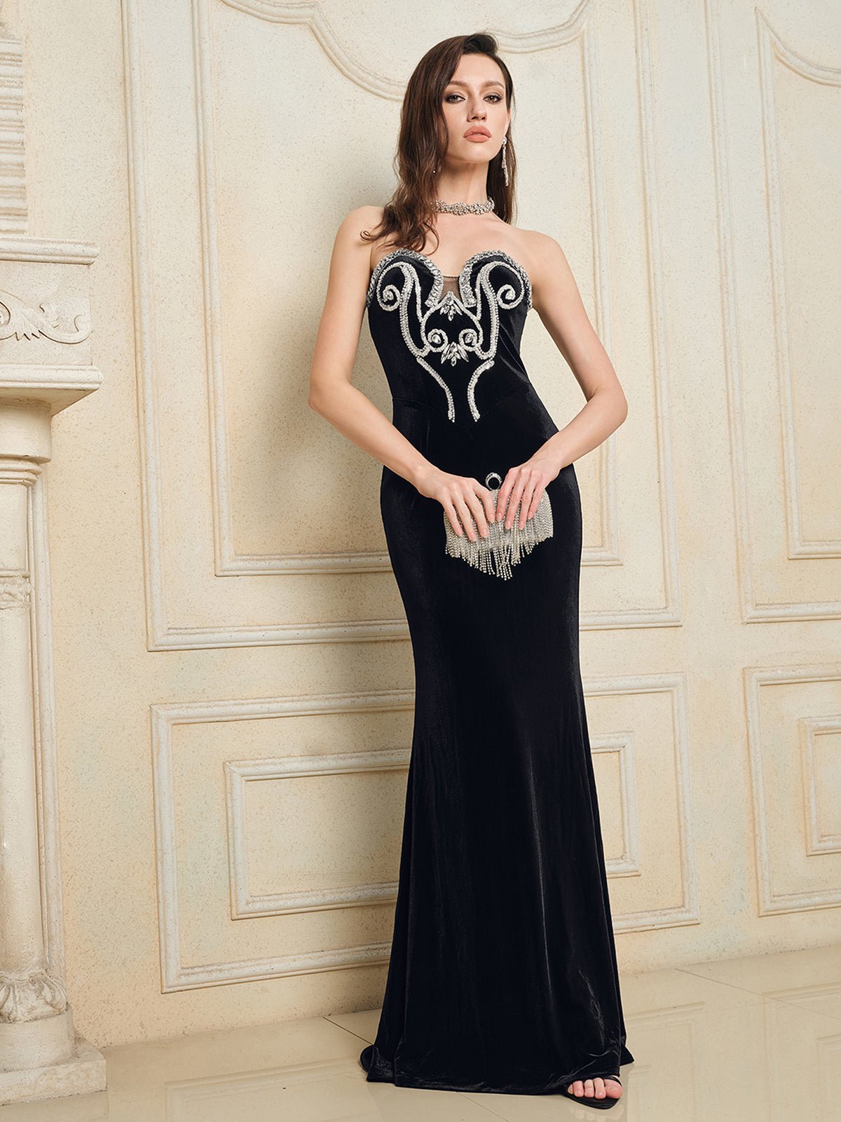A woman wears the Social Queen Hand-Applied Silver Crystal Baroque Velvet Gown, featuring a fitted strapless design with hand-beaded silver crystal swirl accents, paired with a silver fringe clutch and necklace, indoors.
