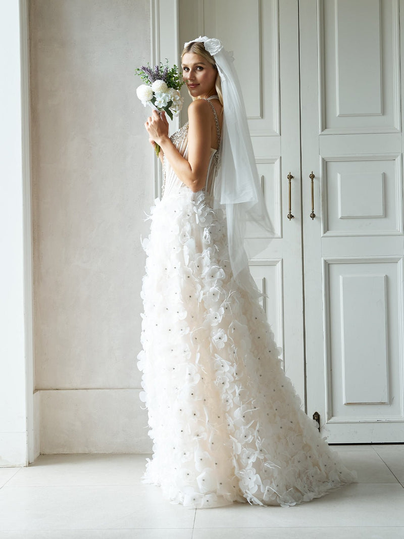A bride in the Social Queen Hand-Beaded 3D Floral Corset Gown with Veil stands indoors, holding a bouquet and looking over her shoulder as her veil flows gracefully behind her.