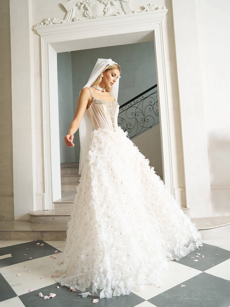 A woman in the Social Queen Hand-Beaded 3D Floral Corset Gown with Veil stands indoors near a staircase, flower petals scattered around her.
