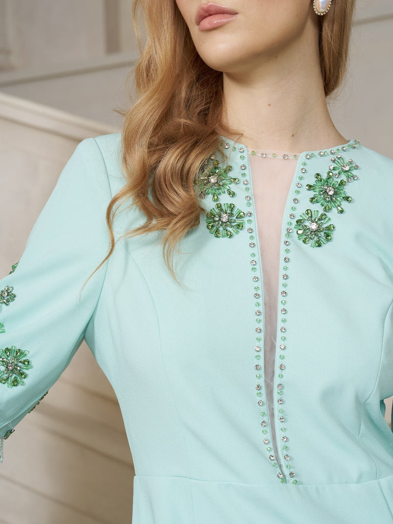 Woman wearing the Social Queen Hand-Beaded Caftan Dress with Fringe Trim, featuring intricate beaded floral embellishments along the neckline and sleeves, standing indoors.