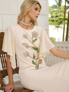 A woman in Social Queen’s Hand-Beaded Floral Mermaid Dress with chiffon cape sleeves and a pearl choker sits outdoors on a wooden chair, gazing to the side.
