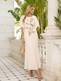 A woman wears the Social Queen Hand-Beaded Floral Mermaid Dress, featuring a long cream design with chiffon cape sleeves, standing on a tiled balcony with white columns and lush greenery behind her.