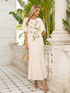 A woman wears the Social Queen Hand-Beaded Floral Mermaid Dress, featuring a long cream design with chiffon cape sleeves, standing on a tiled balcony with white columns and lush greenery behind her.