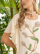 A woman in the Social Queen Hand-Beaded Floral Mermaid Dress, featuring chiffon cape sleeves, a pearl choker, and earrings, stands before lush green plants.