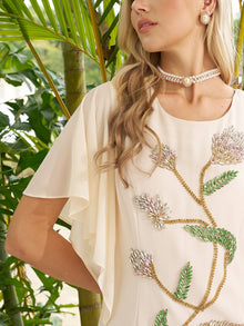 A woman in the Social Queen Hand-Beaded Floral Mermaid Dress, featuring chiffon cape sleeves, a pearl choker, and earrings, stands before lush green plants.