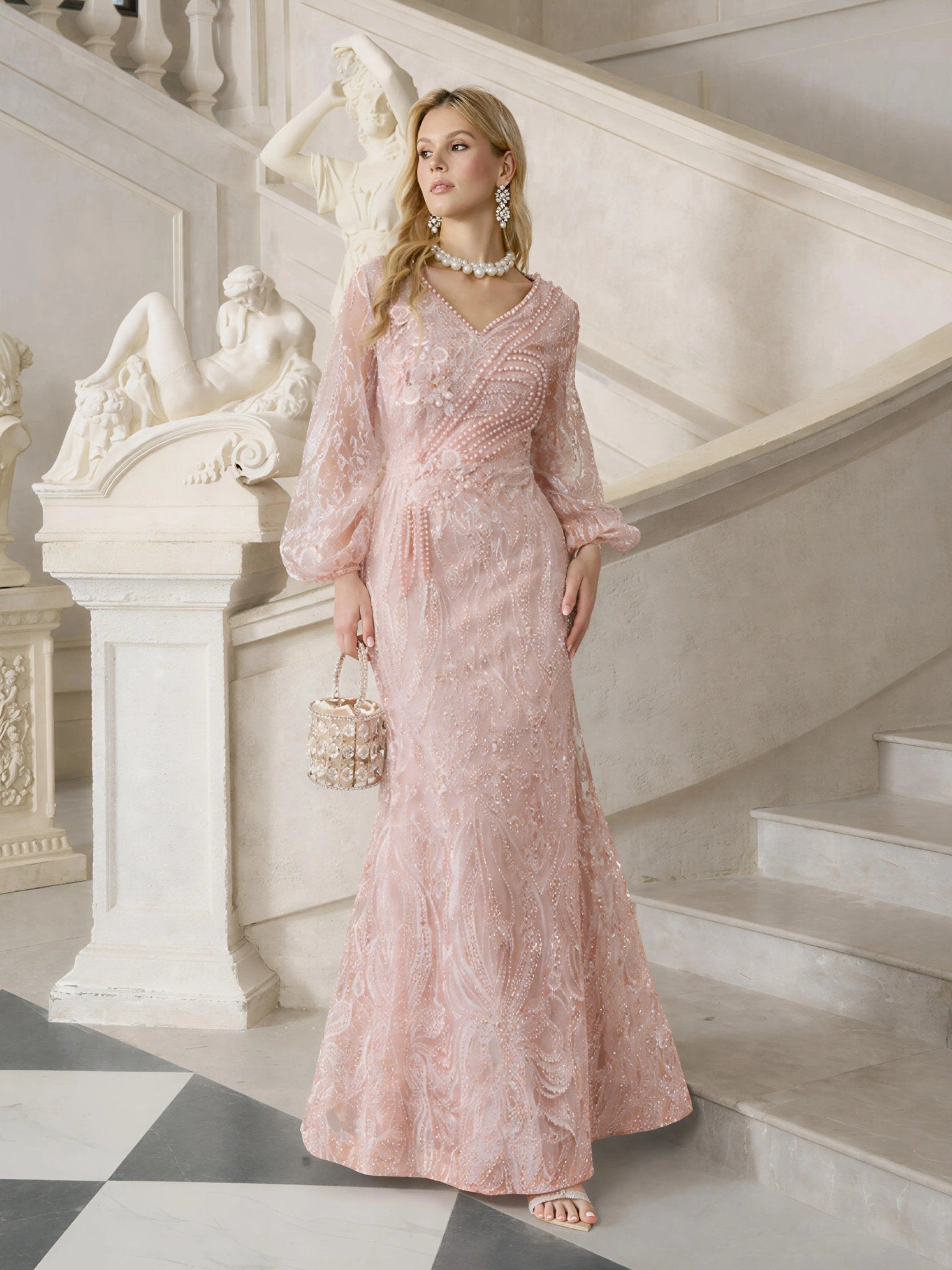 A woman wears the Social Queen Hand-Beaded Lace Mermaid Dress in pink on marble stairs, holding a small embellished handbag, with classical statues in the background.