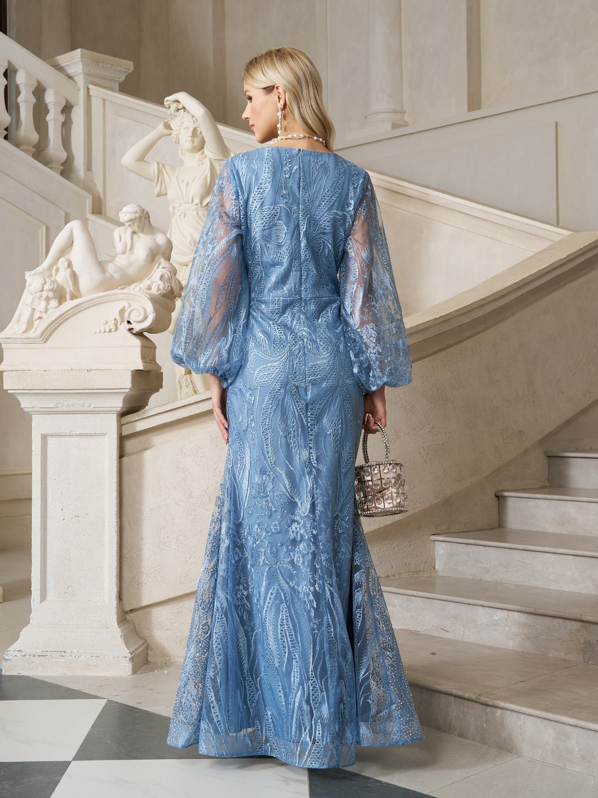 A woman wearing the Social Queen Hand-Beaded Lace Mermaid Dress stands on marble stairs, holding a small handbag, with a classical statue in the background. The gown features sheer sleeves and delicate beaded embellishments.