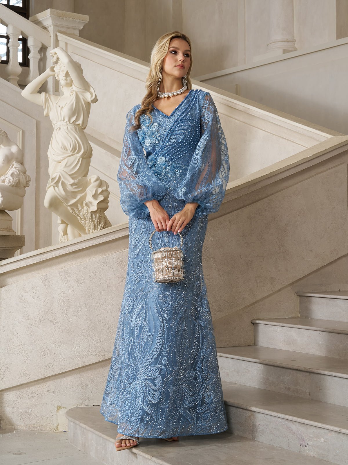 A woman stands on a staircase holding a small handbag, wearing the Social Queen Hand-Beaded Lace Mermaid Dress in light blue with sheer sleeves. A white statue is visible in the background.