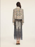 A person stands facing away on a plain light background, wearing the Hand-Beaded Printed Jacket by Social Queen paired with a metallic, fringed skirt and high heels.