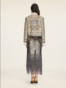 A person stands facing away on a plain light background, wearing the Hand-Beaded Printed Jacket by Social Queen paired with a metallic, fringed skirt and high heels.