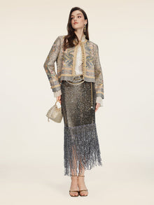A woman stands against a plain background wearing the Social Queen Hand-Beaded Printed Jacket, paired with a white blouse, metallic fringe skirt, gold heels, and a small gold handbag.