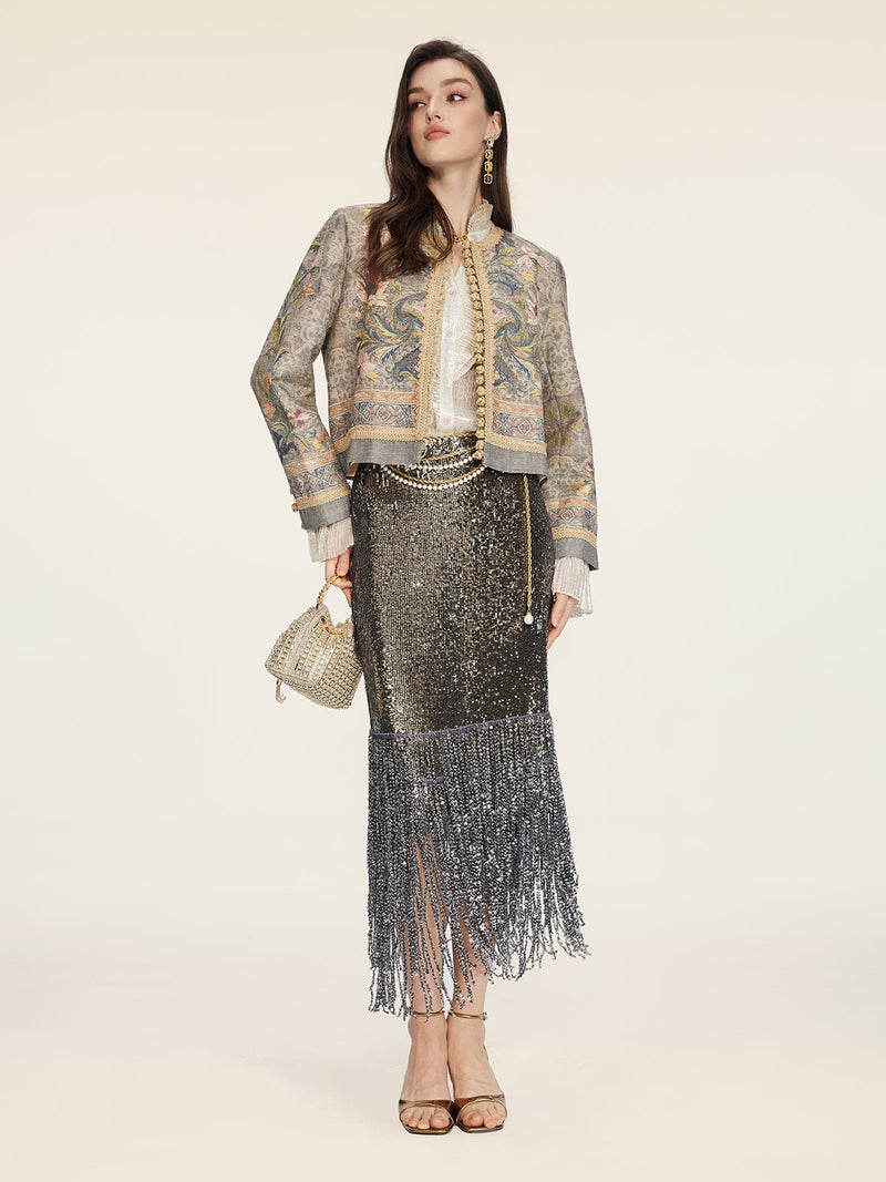 A woman stands against a plain background wearing the Social Queen Hand-Beaded Printed Jacket, paired with a white blouse, metallic fringe skirt, gold heels, and a small gold handbag.