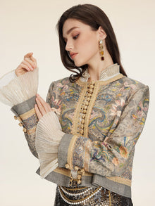 A woman models the Social Queen Hand-Beaded Printed Jacket, featuring floral embroidery, beaded frog closures, gold trim, and pleated cuffs, paired with dangling earrings against a neutral background.