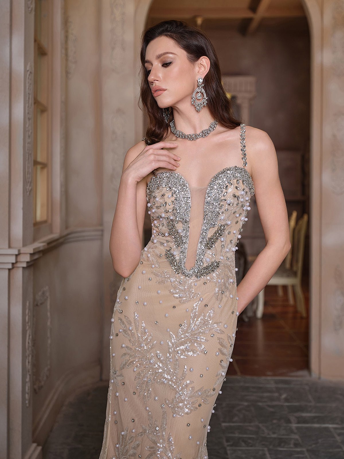 A woman stands indoors wearing the Social Queen Hand-Beaded Sequin Embroidered Cocktail Gown in beige, paired with matching earrings and a necklace, with one hand near her neck.