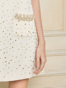A close-up of the Hand-Beaded Silk Wool Embossed Dress by Social Queen, cream-colored tweed with gold and pearl embellishments and a pearl-detailed pocket. A person's arm and hand rest at their side.