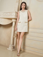 A woman in Social Queen’s Hand-Beaded Silk Wool Embossed Dress stands by a decorated cream-colored wall, holding a gold handbag and wearing gold sandals.