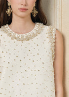 A woman models the Hand-Beaded Silk Wool Embossed Dress by Social Queen, a sleeveless cream A-line style with pearl trim, paired with ornate gold earrings. Only the lower half of her face is visible.