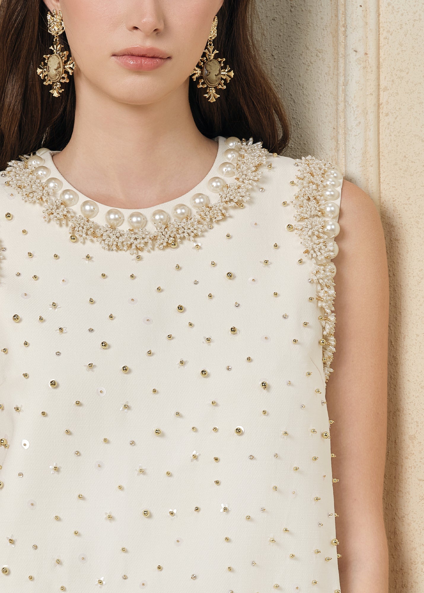 A woman models the Hand-Beaded Silk Wool Embossed Dress by Social Queen, a sleeveless cream A-line style with pearl trim, paired with ornate gold earrings. Only the lower half of her face is visible.