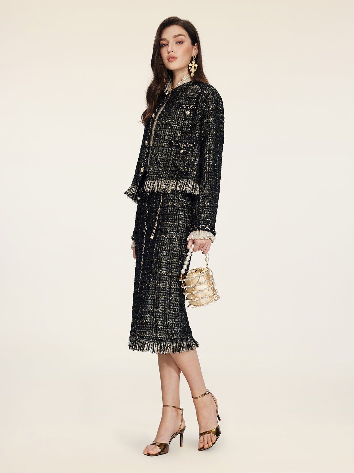 A woman wears the Social Queen Hand-Beaded Tweed Fringe Jacket, styled with a dark skirt suit, gold earrings, metallic heels, and a small gold handbag, standing against a plain light background.