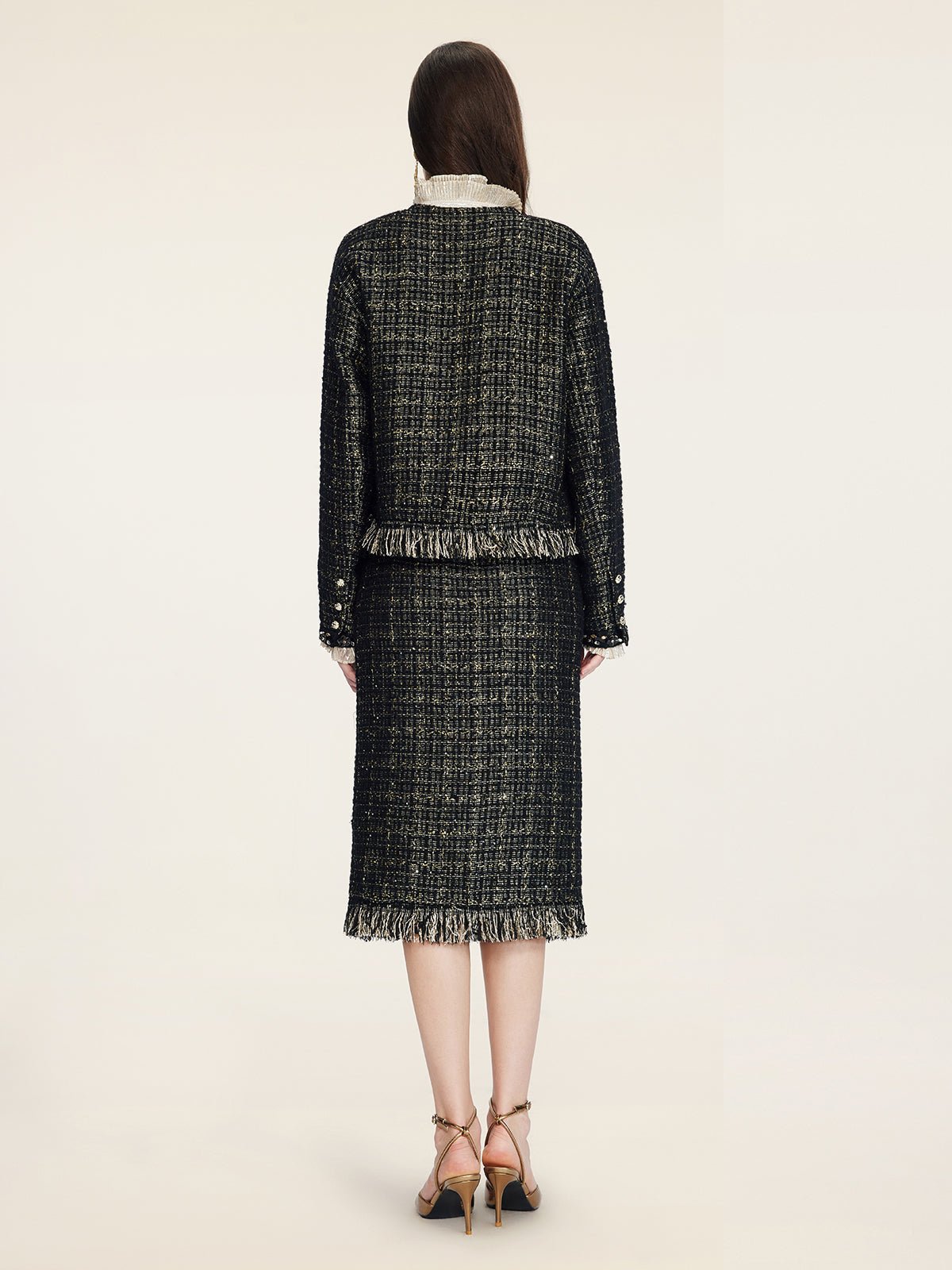 A woman faces away, wearing the Social Queen Hand-Beaded Tweed Fringe Jacket with matching skirt, featuring a cropped silhouette and fringe details, paired with tan heels against a plain light background.