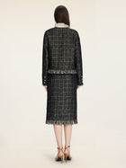 A woman faces away, wearing the Social Queen Hand-Beaded Tweed Fringe Jacket with matching skirt, featuring a cropped silhouette and fringe details, paired with tan heels against a plain light background.