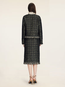 A woman faces away, wearing the Social Queen Hand-Beaded Tweed Fringe Jacket with matching skirt, featuring a cropped silhouette and fringe details, paired with tan heels against a plain light background.