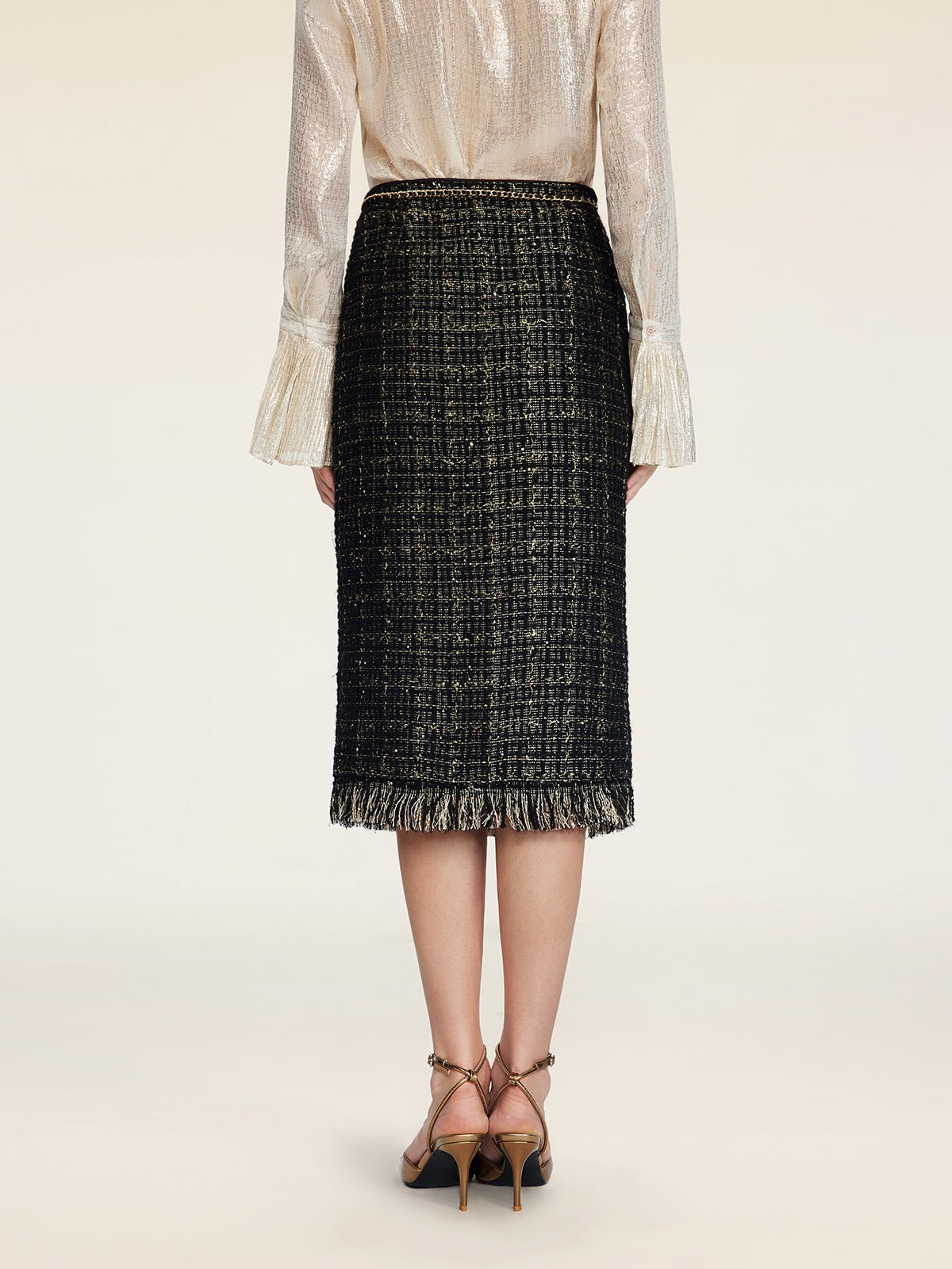 A person stands facing away, wearing a shimmery long-sleeve blouse and the Social Queen Hand-Beaded Tweed Fringe Slit Skirt, featuring black-and-gold tweed with a fringed hem, paired with strappy heels.