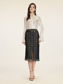 A woman faces forward wearing a sheer white ruffled blouse and the Social Queen Hand-Beaded Tweed Fringe Slit Skirt, featuring black-and-gold details, front buttons, a fringe hem, and paired with strappy heeled sandals.