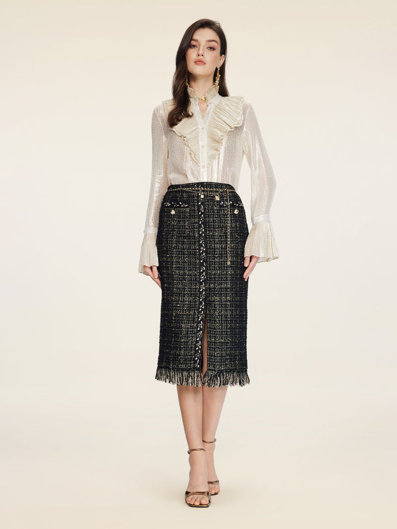 A woman faces forward wearing a sheer white ruffled blouse and the Social Queen Hand-Beaded Tweed Fringe Slit Skirt, featuring black-and-gold details, front buttons, a fringe hem, and paired with strappy heeled sandals.