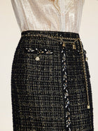 A person wears the Social Queen Hand-Beaded Tweed Fringe Slit Skirt, featuring black-and-gold tweed, decorative chains, floral embellishments, front buttons, and intricate hand-applied beadwork.