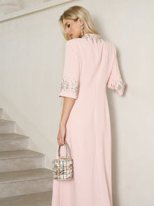 A woman wearing the Social Queen Hand-Embellished A-Line Dress in light pink, featuring beaded sleeves, stands by a staircase holding a silver handbag, facing away from the camera.