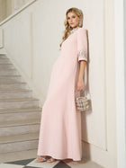 A woman in a pale pink, floor-length dress with embellished sleeves stands on a staircase, holding a silver handbag and wearing silver sandals and statement earrings.