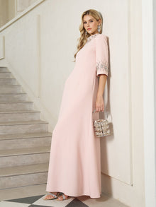 A woman in a pale pink, floor-length dress with embellished sleeves stands on a staircase, holding a silver handbag and wearing silver sandals and statement earrings.