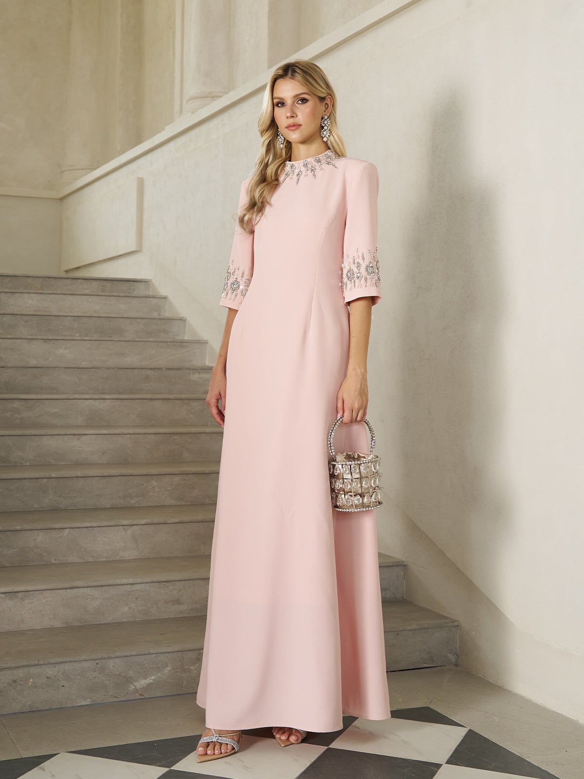 A woman stands on stone stairs holding a small handbag, wearing the Social Queen Hand-Embellished A-Line Dress in pale pink, floor-length with half sleeves.
