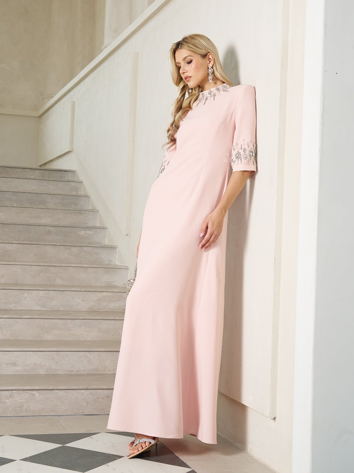 A woman wearing the Social Queen Hand-Embellished A-Line Dress in light pink stands indoors by a staircase, showcasing its floor-length silhouette and silver details.