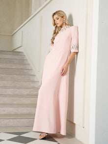 A woman wearing the Social Queen Hand-Embellished A-Line Dress in light pink stands indoors by a staircase, showcasing its floor-length silhouette and silver details.