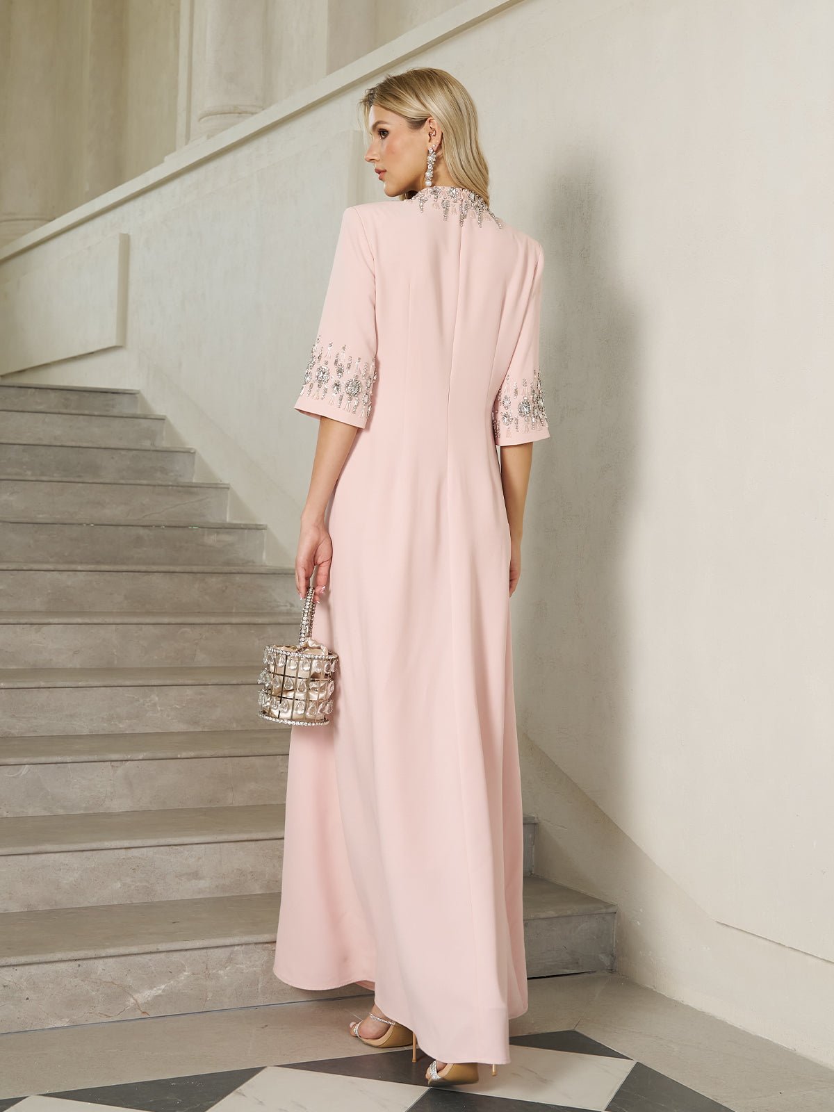A woman in the Social Queen Hand-Embellished A-Line Dress, featuring a long pale pink silhouette, stands on marble stairs with a small square handbag, facing away from the camera.
