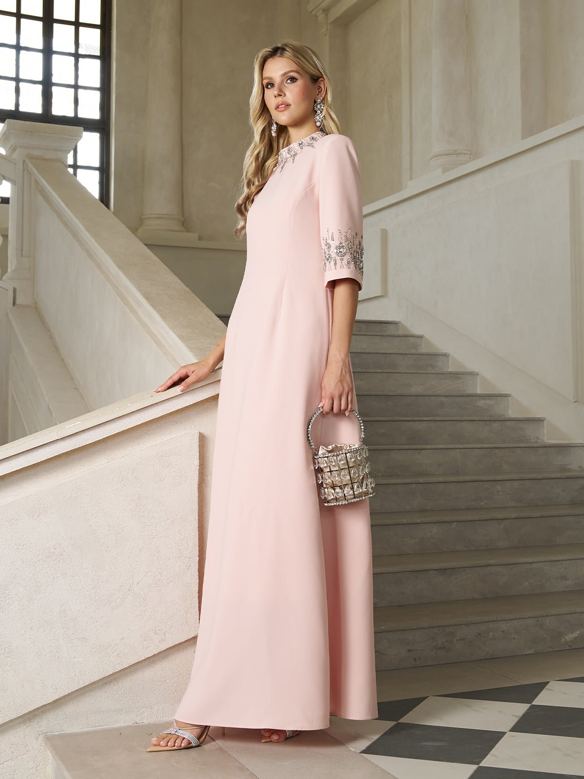 A woman in the Social Queen Hand-Embellished A-Line Dress, featuring embellished sleeves and a pale pink hue, stands on a staircase holding a matching embellished handbag—a refined wedding guest look.