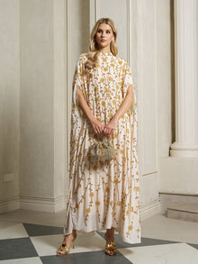 A woman stands indoors wearing the Social Queen Hand-Embellished Floral Caftan Dress with gold heels and a small embellished handbag, against a backdrop of pillars and a checkered floor for an elegant resort wear look.
