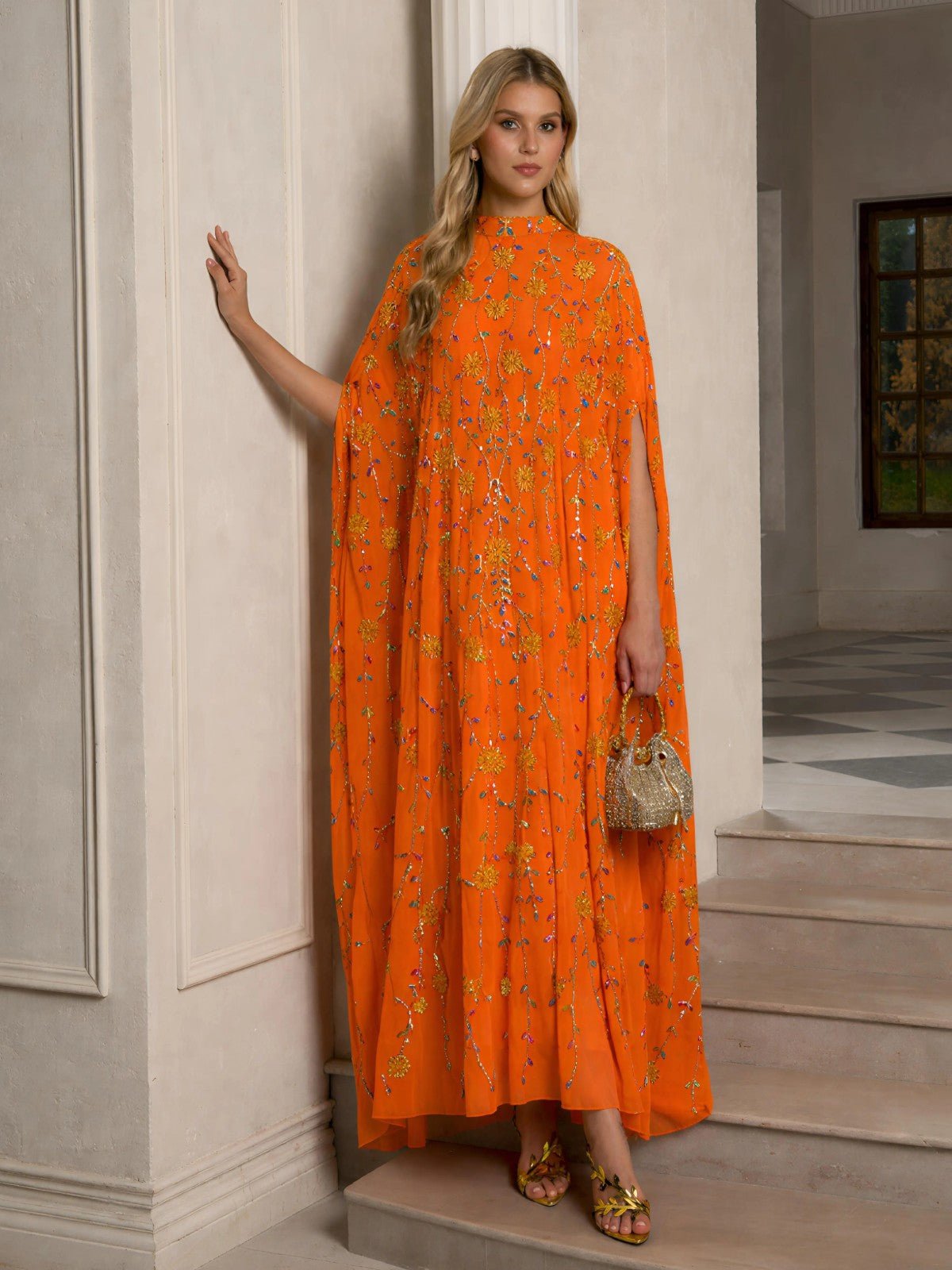 A woman indoors wears the Social Queen Hand-Embellished Floral Caftan Dress with a cape design, paired with a beaded handbag and gold sandals—an ideal choice for chic resort style.