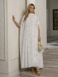 A woman stands indoors in the Hand-Embellished Floral Caftan Dress by Social Queen—a long, white floral piece with delicate embroidery and a cape—styled with a beaded handbag and gold sandals for resort elegance against neutral walls and a window.