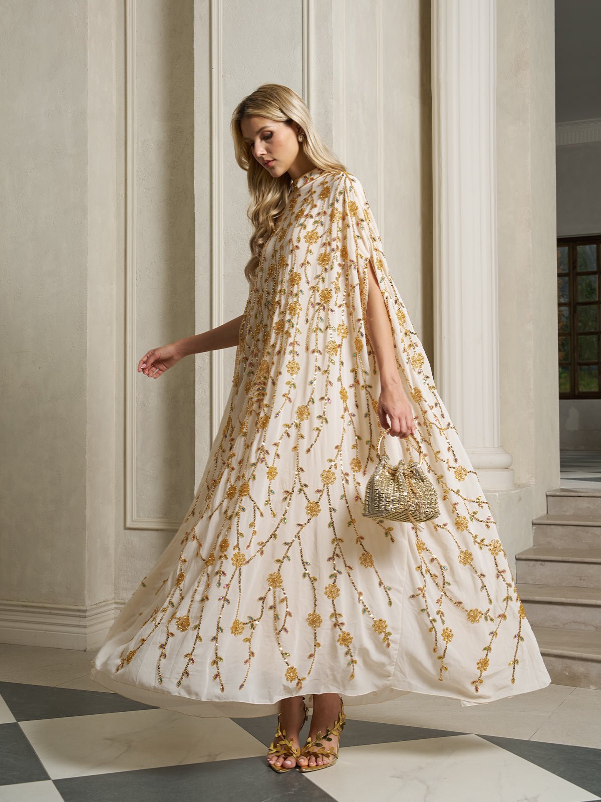 A woman wears the Social Queen Hand-Embellished Floral Caftan Dress, featuring gold embroidery, paired with a gold handbag and matching sandals—an elegant choice for resort wear.
