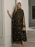 Indoors, a woman wears the Social Queen Hand-Embellished Floral Caftan Dress with gold patterns, paired with gold sandals and a small gold handbag.