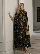 Indoors, a woman wears the Social Queen Hand-Embellished Floral Caftan Dress with gold patterns, paired with gold sandals and a small gold handbag.