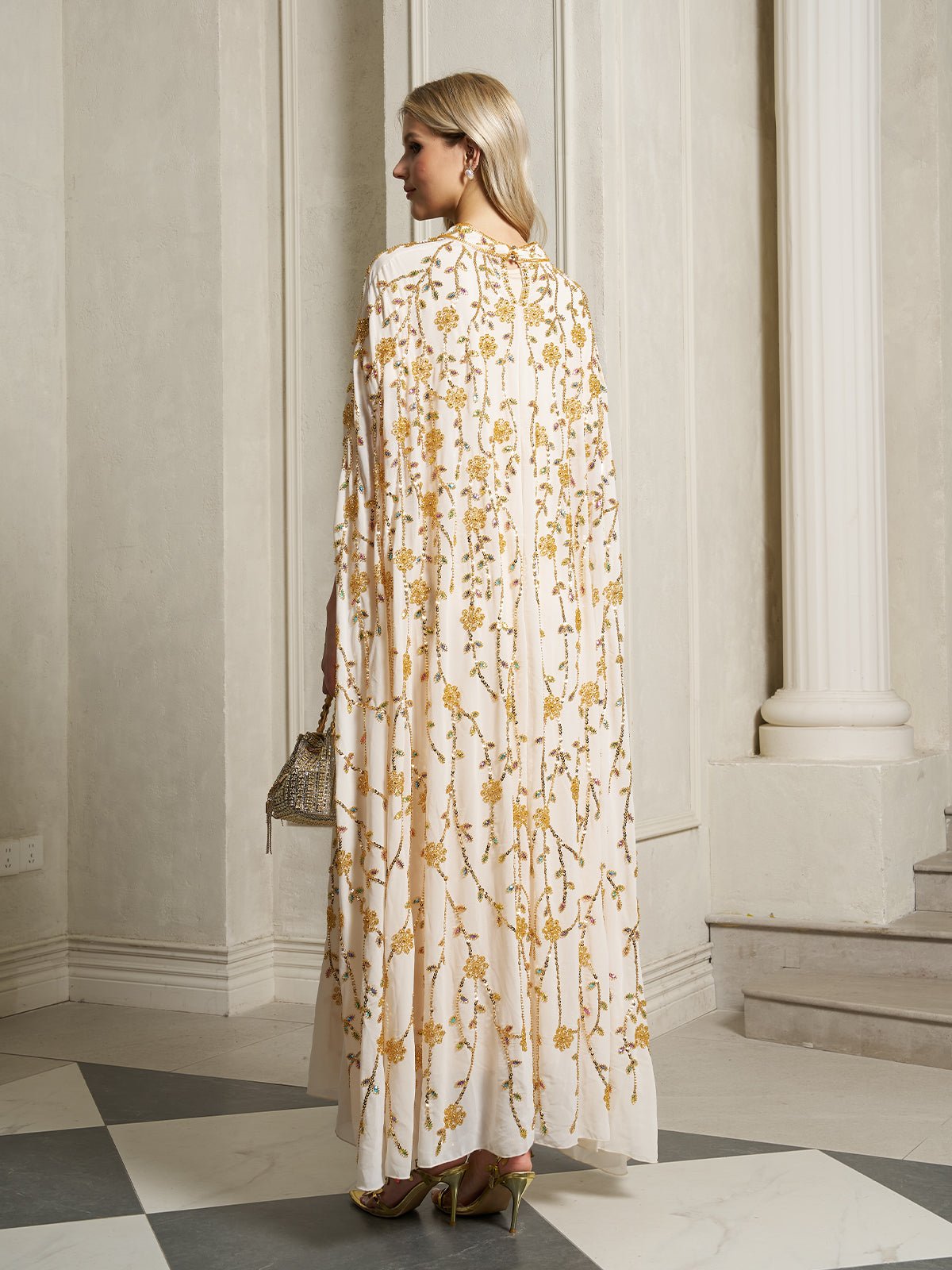 A woman stands indoors with her back to the camera, holding a small handbag and wearing the Social Queen Hand-Embellished Floral Caftan Dress in a floor-length white and gold design.