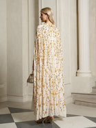 A woman stands indoors with her back to the camera, holding a small handbag and wearing the Social Queen Hand-Embellished Floral Caftan Dress in a floor-length white and gold design.