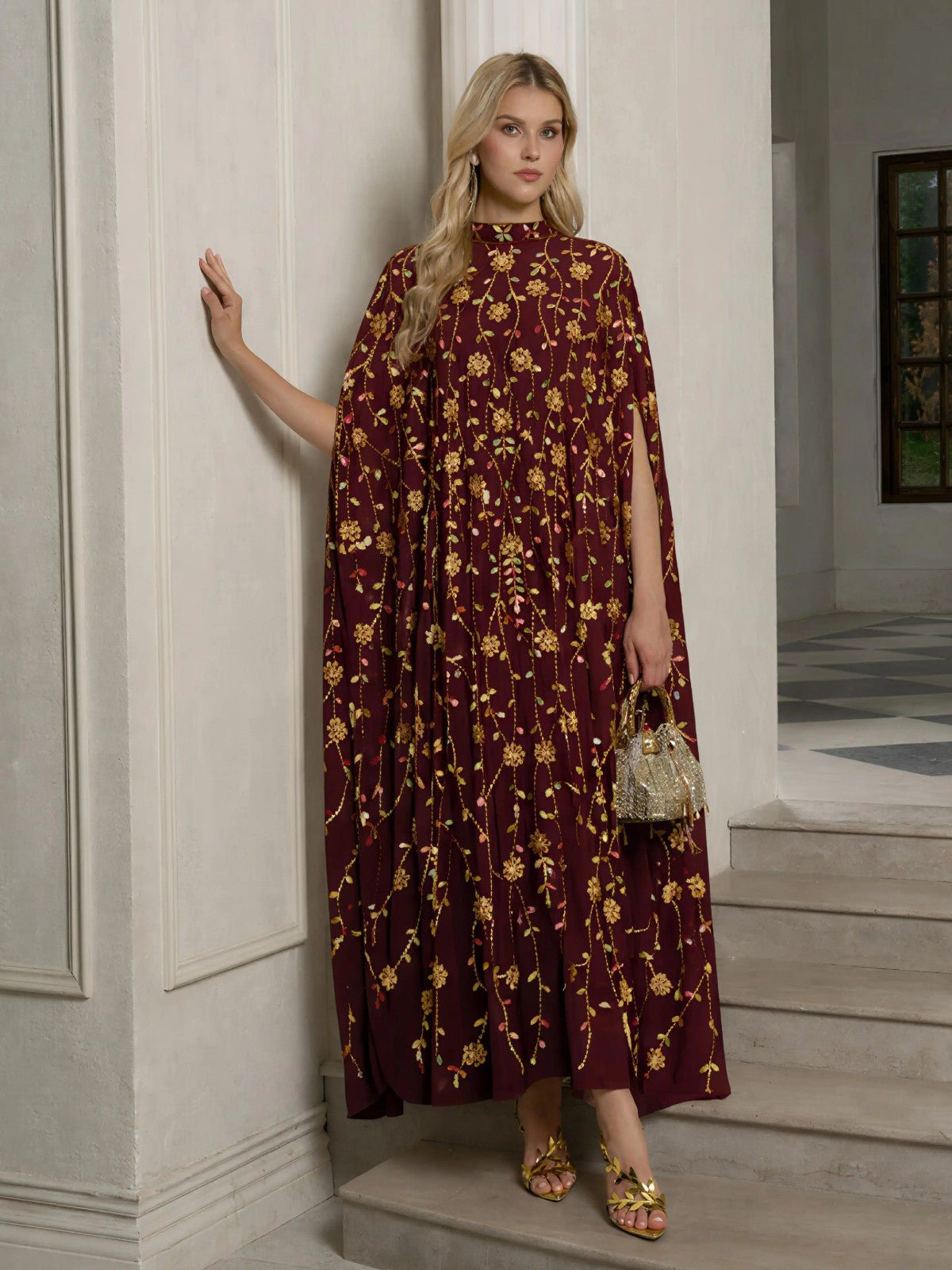 A woman stands in a room wearing the Social Queen Hand-Embellished Floral Caftan Dress, paired with gold sandals and holding a small gold handbag.