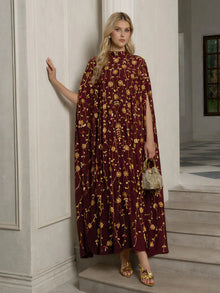 A woman stands in a room wearing the Social Queen Hand-Embellished Floral Caftan Dress, paired with gold sandals and holding a small gold handbag.