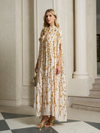 A woman stands indoors wearing the Social Queen Hand-Embellished Floral Caftan Dress, featuring gold embroidery. She pairs it with gold sandals and a small gold handbag, adding elegant resort wear flair against marble floors and columns.